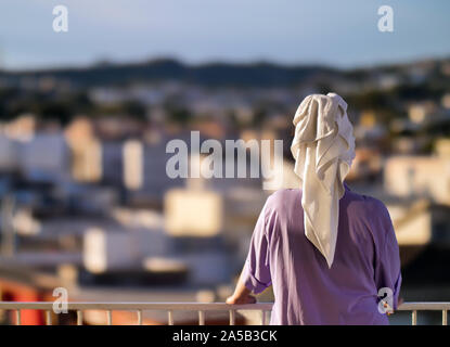 Una donna si erge su un balcone al mattino con un asciugamano bianco sulla sua testa contro lo sfondo della città, vista posteriore Foto Stock