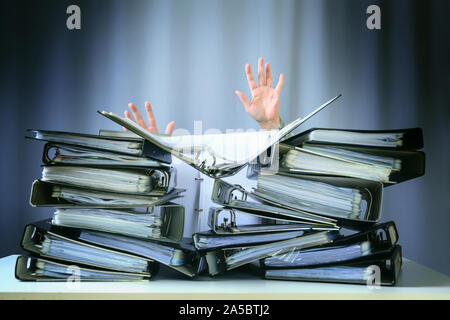 Alzare le mani di una persona che affonda dietro pile di raccoglitori ad anello su una scrivania in ufficio, concetto di richieste eccessive e aumentando il lavoro in azienda, sele Foto Stock