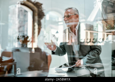 Il brainstorming. Grave uomo maturo in abbigliamento formale per spiegare qualcosa con i colleghi e gesticolando mentre è seduto al tavolo per ufficio dietro Foto Stock