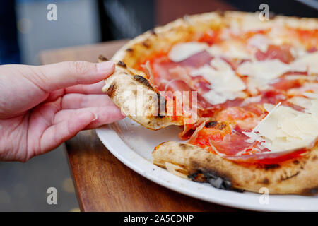 Mano pezzo di prelevamento della tradizionale Prosciutto di Parma la pizza fatta con il classico forno a legna, sulla parte superiore con formaggio parmigiano, servire su piastra bianca e un tavolo di legno. Foto Stock