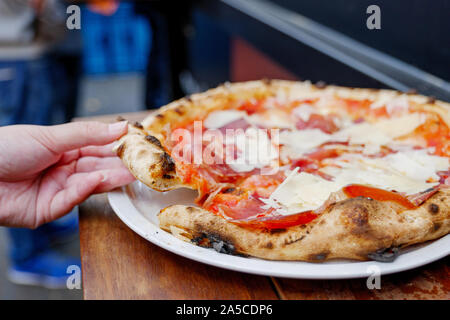 Mano pezzo di prelevamento della tradizionale Prosciutto di Parma la pizza fatta con il classico forno a legna, sulla parte superiore con formaggio parmigiano, servire su piastra bianca e un tavolo di legno. Foto Stock