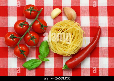 Materie prime: spaghetti, pomodori ciliegini, peperoncino rosso, spicchi di aglio e di foglie di basilico fresco su rosso e bianco di panno a scacchi Foto Stock