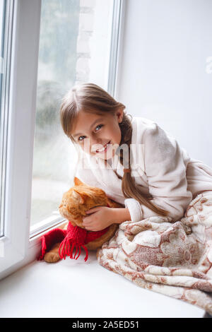 Bambina in abito teddy seduta su una soglia finestra con animale domestico. Weekend pigro con gatto rosso a casa. Foto Stock