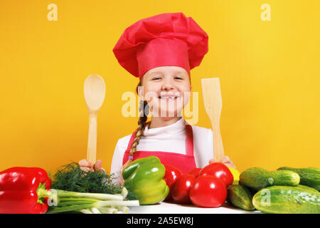 Bellissima bambina chef su sfondo giallo. Il bambino si siede a un tavolo con verdure e tiene nelle sue mani un cucchiaio di legno e una spatola. Foto Stock