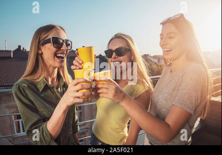 Merry ragazze in occhiali da sole toccando bicchieri su un balcone in raggi del sole al tramonto Foto Stock