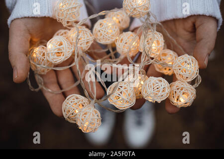 Luci del Palms. Mani femminili tenere una ghirlanda di vimini con le sfere rotonde. Ragazza in un maglione bianco con le luci di Natale nelle sue mani, caldo nuovo anno Foto Stock