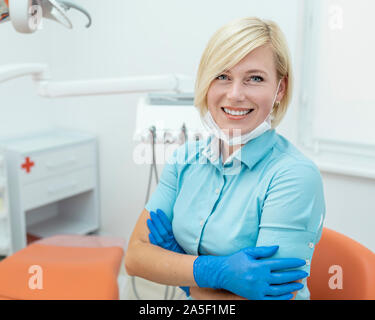 Ritratto di sorridente femmina bionda dentista in blu scrubs e guanti Foto Stock
