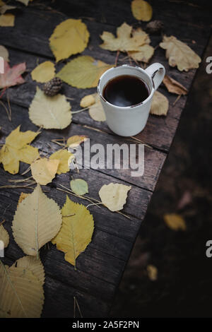 Su un vecchio tavolo di legno in autunno park è una tazza di tè e caffè, sparse in foglie di giallo e pigne. Vista dall'alto, in blur. Autunno Caldo buio Foto Stock