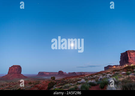 Formazioni di Butte un ampio angolo di visione nella Monument Valley durante il crepuscolo blu notte oscura in Arizona con vetture su polvere di strada sterrata e la luna nel cielo Foto Stock