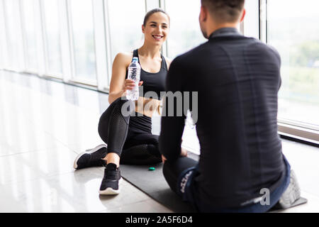 Stanco di idoneità giovane avente il resto e parlare, acqua potabile in palestra. L uomo e la donna seduta sul pavimento, rottura dopo la formazione, uno stile di vita sano concetto Foto Stock