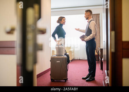 Piuttosto giovane donna elegante con i bagagli in piedi nella camera di albergo e parlando a porter accogliendo il suo Foto Stock