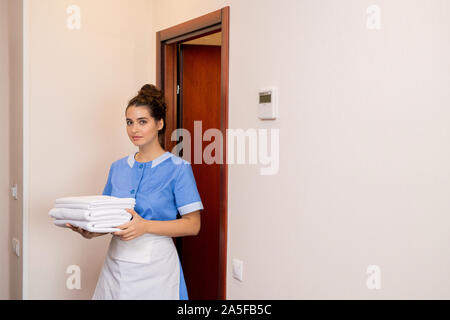 Bruna giovane cameriera in uniforme che porta pila di bianco asciugamani freschi mentre andando fuori di una delle camere Foto Stock