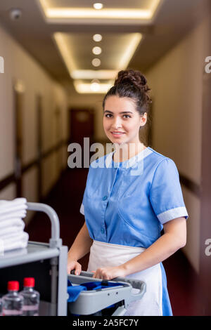 Giovani sorridente brunette femmina in uniforme blu guardando a voi mentre si muove lungo il corridoio in hotel e spingendo il carrello con roba di igiene Foto Stock