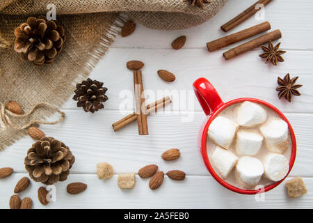 Natale rosso con tazza di cioccolata calda e marshmallow. Cioccolata calda con latte, anice e bastoncini di cannella, mandorle e pigne su bianco rustico in legno ba Foto Stock