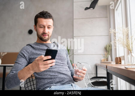 Giovane uomo riposante rilassante in intimo cafe, avente un bicchiere di acqua e di scorrimento o di messaggistica dello smartphone in Foto Stock