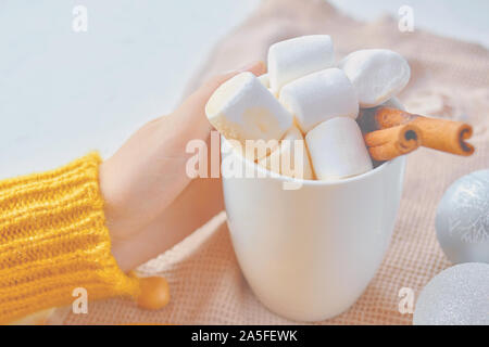 Donne mano, maglione giallo e bianco mug. Con una bevanda calda, marshmallow e la stecca di cannella Foto Stock