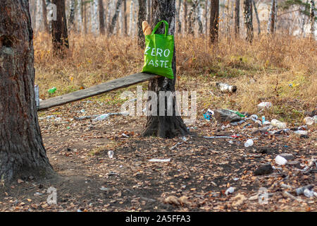 Un tessile borsa verde con la scritta rifiuti zero sorge nella foresta accanto alla spazzatura sparsi sul terreno. Un sacco di piatti di plastica e bott Foto Stock