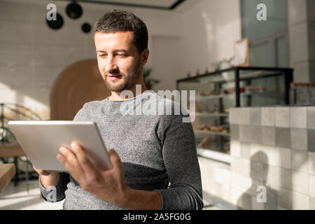 Felice giovane con gadget mobile in piedi in camera da sole o cafe durante lo scorrimento attraverso la roba online Foto Stock