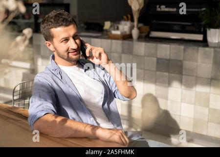 Felice giovane impreditore con lo smartphone parlando a un collega o client di consulenza durante i momenti di relax da tavola in cafe Foto Stock