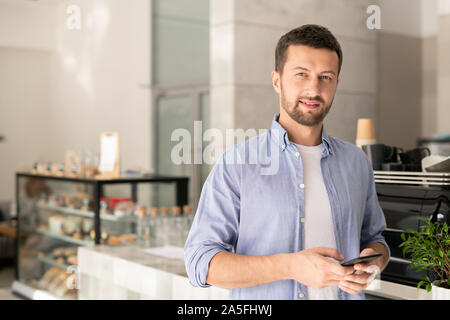 Bel ragazzo in informale lo scorrimento in smartphone mentre ti guarda sullo sfondo del Cafe interior Foto Stock