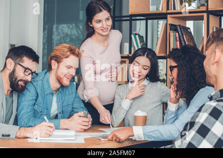 Startupers lavorando insieme a Office seduta guardando i documenti allegro mentre la donna incinta in piedi rivolta a idea per il progetto Foto Stock