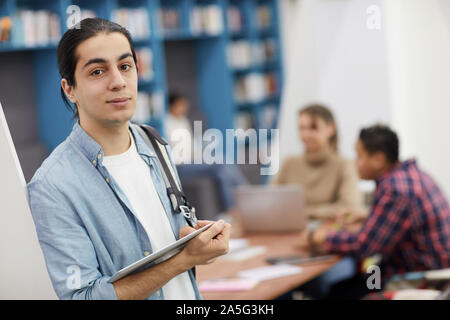 Vita ritratto di con i capelli lunghi uno studente universitario che guarda la fotocamera mentre in piedi in biblioteca moderna, spazio di copia Foto Stock
