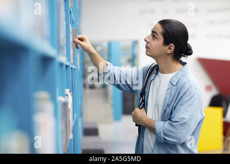 Vista laterale ritratto di studente di Latino-americano la scelta di libri in piedi da ripiani in college biblioteca, spazio di copia Foto Stock