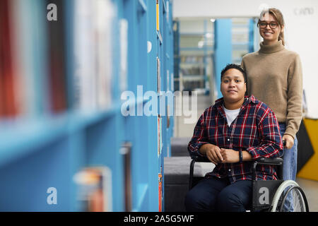 Ritratto di due studenti del college, uno di essi in carrozzella, che pongono insieme in piedi da ripiani in biblioteca, spazio di copia Foto Stock