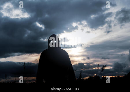 Un concetto di orrore. Una silhouette di una figura con cappuccio, stando in piedi in un campo , con paura gli occhi luccicanti guardando la telecamera. Retro illuminato dall'impostazione s Foto Stock