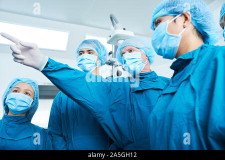 Senior chirurgo in scrub, la maschera per il viso e il tappo rivolto al monitor di visualizzare informazioni circa lo stato di salute del paziente al gruppo di giovani stagisti Foto Stock