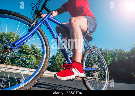 Montare maschio ciclista biker in sella alla sua moto ciclo su una strada asfaltata al tramonto mentre il sole tramonta attraverso la sua ruota. Foto Stock