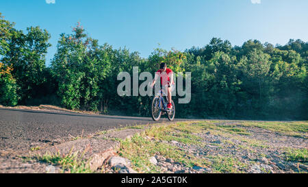 Montare maschio ciclista biker in sella alla sua moto ciclo su una strada asfaltata al tramonto mentre il sole tramonta attraverso la sua ruota. Foto Stock