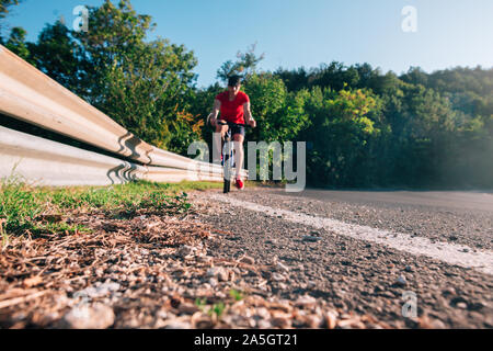 Montare maschio ciclista biker in sella alla sua moto ciclo su una strada asfaltata al tramonto mentre il sole tramonta attraverso la sua ruota. Foto Stock