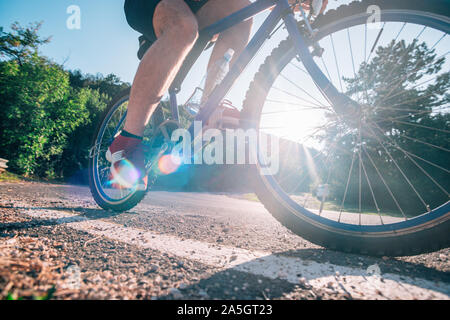 Montare maschio ciclista biker in sella alla sua moto ciclo su una strada asfaltata al tramonto mentre il sole tramonta attraverso la sua ruota. Foto Stock