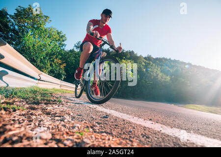 Montare maschio ciclista biker in sella alla sua moto ciclo su una strada asfaltata al tramonto mentre il sole tramonta attraverso la sua ruota. Foto Stock