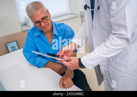 Persone di mezza età caucasian dottore spiega al suo paziente senior del suo sangue le letture della pressione mentre in piedi accanto a lui. Foto Stock