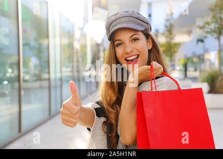 Sorridenti moda donna azienda shopping bag nella sua mano e dà il pollice fino a telecamera in un centro commerciale per lo shopping. Foto Stock