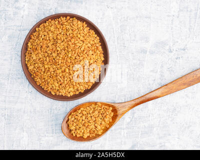 Spezie ed erbe contribuiscono a mantenere in buona salute e a migliorare l'appetito, vista dall'alto su sfondo di calcestruzzo. Fenugreek nella piastra di argilla e il cucchiaio di legno. Moderno Foto Stock