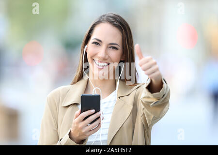 Vista frontale il ritratto di una ragazza felici ascoltando la musica che indossa gli auricolari e azienda smart phone in posa con il pollice su per la strada Foto Stock