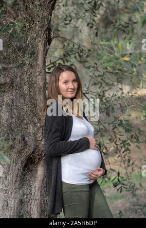 Ritratto di sorridere donna incinta mentre tiene il suo ventre mentre appoggiata contro un albero di olivo Foto Stock