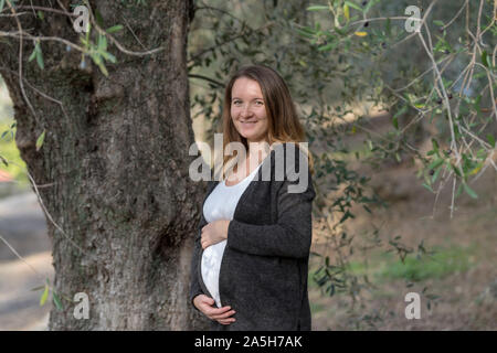 Ritratto di sorridere donna incinta mentre tiene il suo ventre mentre appoggiata contro un albero di olivo Foto Stock