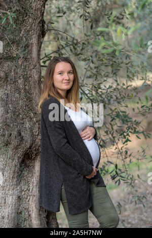 Ritratto di sorridere donna incinta mentre tiene il suo ventre mentre appoggiata contro un albero di olivo Foto Stock