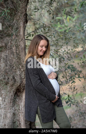 Ritratto di sorridere donna incinta mentre tiene il suo ventre mentre appoggiata contro un albero di olivo Foto Stock