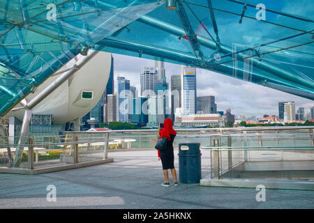 Un giovane turista femminile, coperto con una sciarpa rossa a Ponte di elica, Marina Bay, Singapore, fotografare lo skyline sull'altro lato della baia Foto Stock