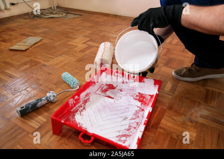 Operaio edile finitura facendo lavori di rinnovo nell'appartamento. Pittore con guanti neri si riversa il colore bianco dalla benna nella vaschetta di colore rosso per la miscelazione della vernice. Foto Stock