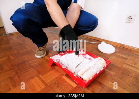 Operaio edile finitura facendo lavori di rinnovo nell'appartamento. Pittore con guanti bianchi mantiene la spazzola a rullo in mano e si mescola la vernice nel contenitore. Foto Stock