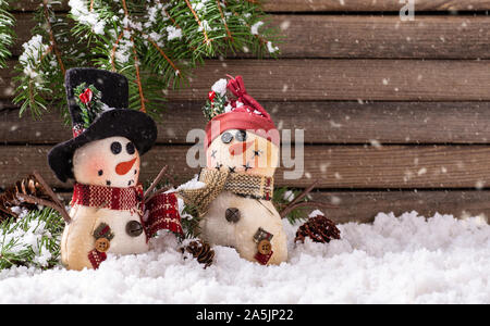Due pupazzi di neve di Natale ornamenti sulla superficie nevosa con caduta di neve su un rustico in legno con sfondo spazio copia Foto Stock