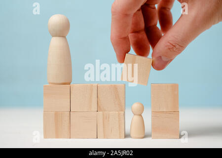 Maschio si chiude a mano in legno di piccole dimensioni in figura dai blocchi sotto la supervisione di un altra figura su sfondo blu. Foto Stock