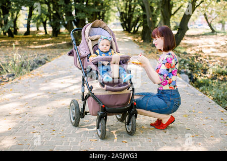 Mamma spoon-alimenta il bambino camminare con il passeggino in posizione di parcheggio . Foto Stock