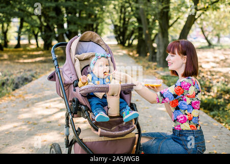 Mom alimentando la sua bambina con un cucchiaio. Madre dando cibo per dieci mesi bambino camminare con il passeggino in posizione di parcheggio . Alimenti per bambini. Foto Stock